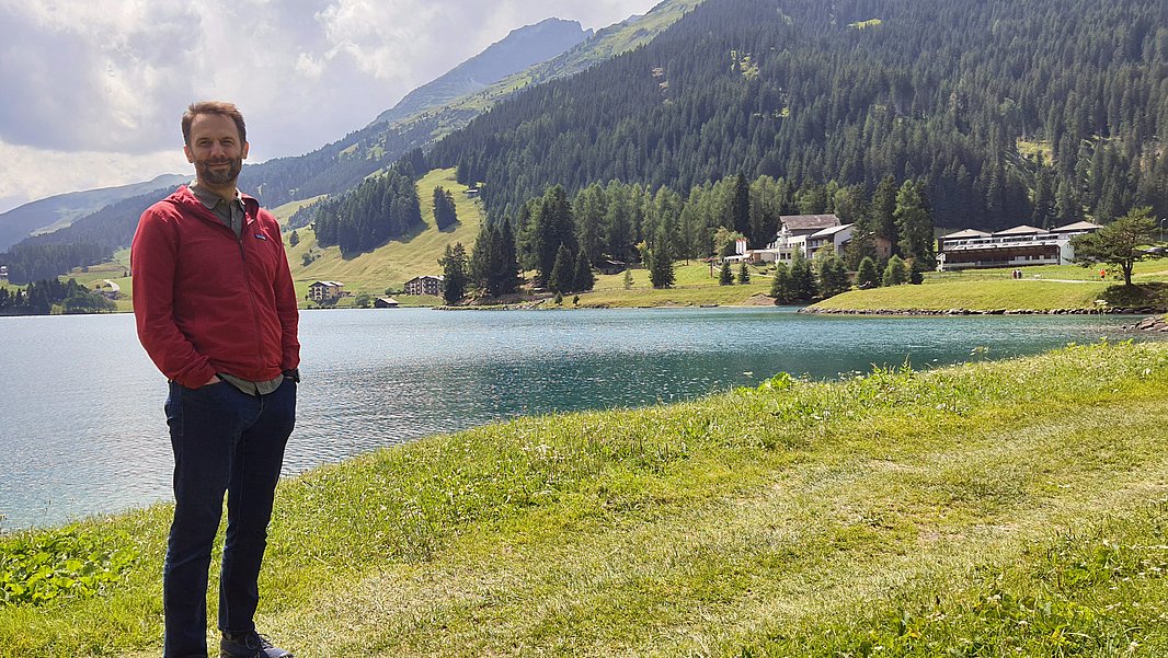 Mann mit Bart in roter Jacke steht vor einem See in einer Gebirgslandschaft.