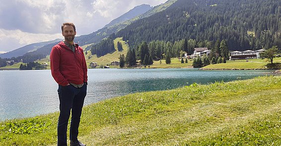 Mann mit Bart in roter Jacke steht vor einem See in einer Gebirgslandschaft.