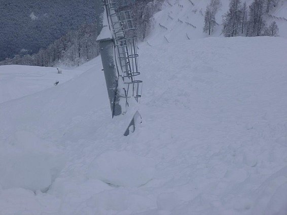 Eine schneebedeckte Landschaft mit einer schräg stehenden Metallstruktur, die vermutlich Teil einer Seilbahn oder eines Skilifts ist. Im Hintergrund sind schneebedeckte Bäume und ein bewölkter Himmel zu sehen.