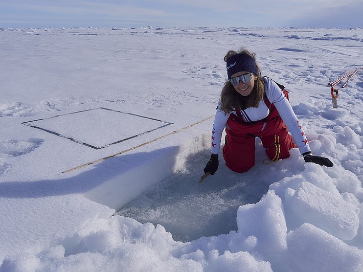 Eine Person kniet auf einer Schneedecke und schaut in ein quadratförmiges Loch im Eis. Sie trägt eine rote Hose und ein weißes Oberteil. Neben ihr liegt ein Zollstock.