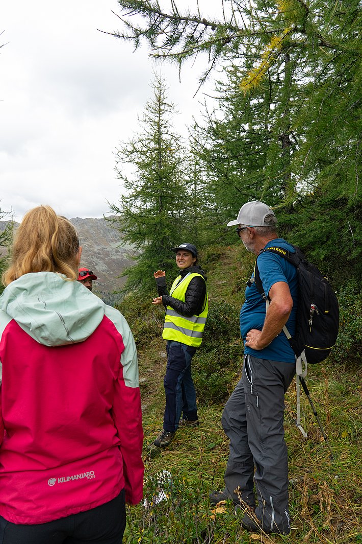 Vier Personen stehen im Freien, umgeben von Bäumen. Eine Person trägt eine gelbe Warnweste. Die anderen Personen sind in Freizeitkleidung gekleidet.