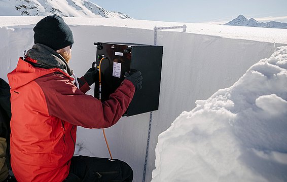 Un uomo con una giacca rossa e un berretto scuro sta posizionando un dispositivo nero all'interno di una grande fessura di neve. Sullo sfondo si possono vedere montagne innevate e un cielo sereno. L'immagine suggerisce un'attività di ricerca o monitoraggio in un ambiente di alta montagna.