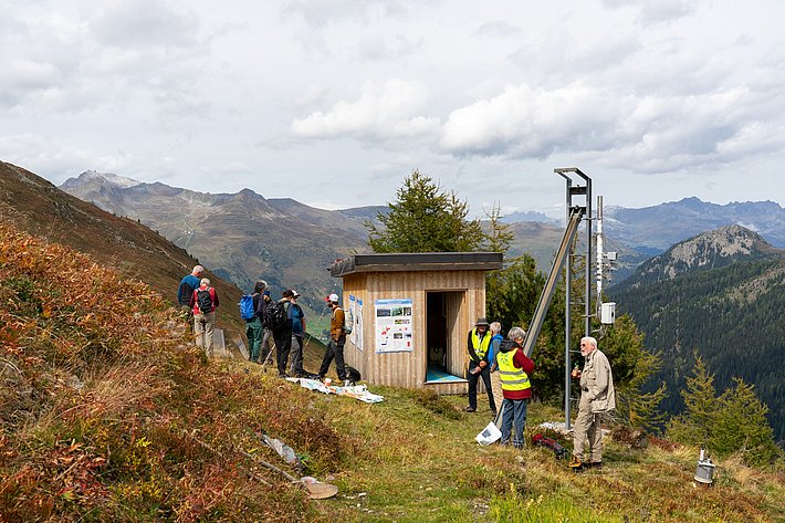 Gruppe von Personen in Wanderkleidung, einige tragen Warnwesten, stehen vor einem kleinen Holzgebäude. Im Hintergrund Berge und bewaldete Hügel.