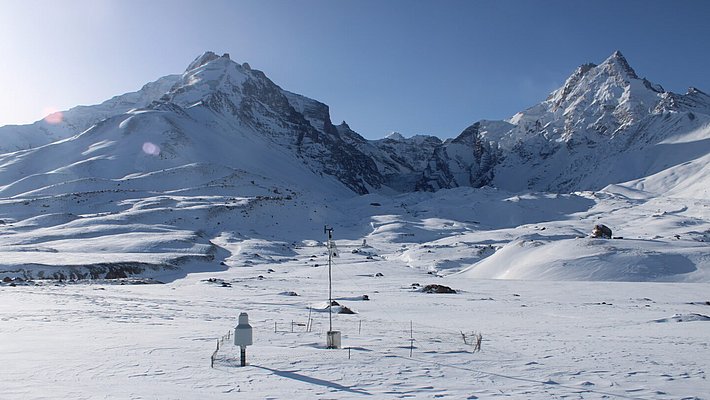 Schneebedeckte Berglandschaft mit klarem blauen Himmel. Im Vordergrund steht eine Wetterstation mit Sensoren, die Schatten auf den unberührten Schnee wirft. Im Hintergrund ragen majestätische Gipfel empor und vermitteln ein Gefühl der Ruhe.