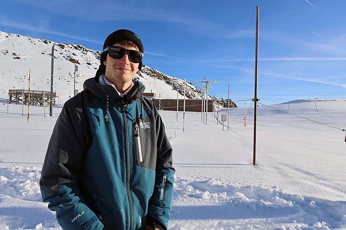 A person wearing a dark jacket and sunglasses stands in a snowy landscape, smiling. They are on a clear day with a backdrop of snow-covered hills and a building visible in the distance. The scene conveys a sense of winter exploration or research.