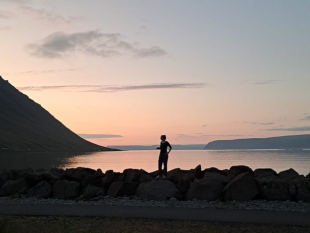 Eine Person steht auf einem Felsen am Ufer eines ruhigen Gewässeres, während die Sonne untergeht und den Himmel in sanften Farben beleuchtet. Im Hintergrund sind sanfte Hügel zu sehen. Die Szene vermittelt Ruhe und natürliche Schönheit.