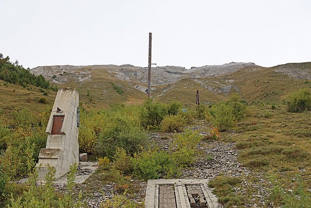 Paysage vallonné avec des montagnes en arrière-plan. Au premier plan, un monument en béton et un poteau, entourés de verdure et de buissons. Le ciel est nuageux et le terrain présente des herbes et des roches.
