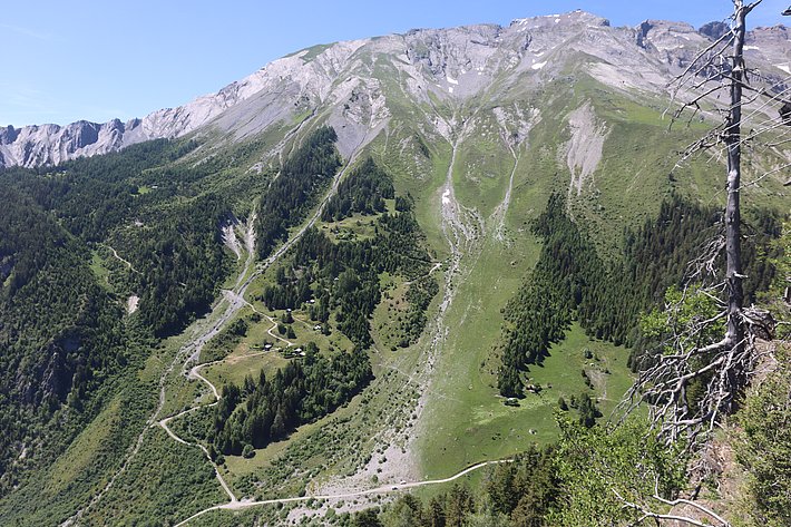 A panoramic view of a mountainous landscape featuring steep slopes, greenery, and rocky terrain under a clear blue sky. Dense forests line the mountain base, while winding paths are visible throughout the area, showcasing the natural beauty of the region.