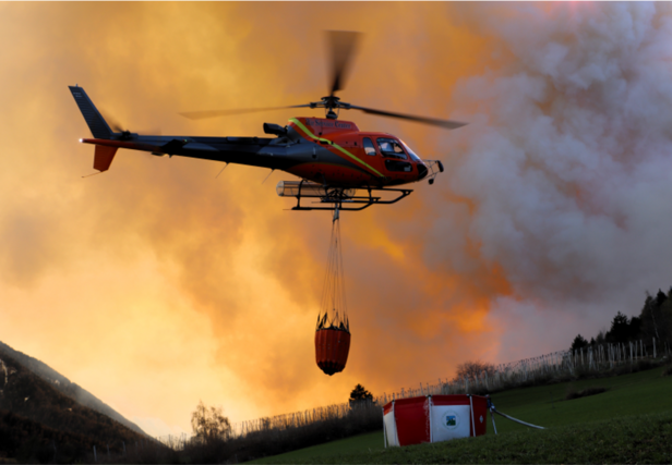 A helicopter carrying a red water tank flies over a green field where there is a tent belonging to the fire brigade, with orange smoke visible in the background.