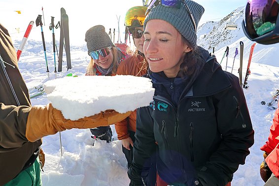 A group of four people in winter clothing is gathered in a snowy landscape. One person is holding a chunk of snow, while another leans in to examine it closely. Ski equipment is visible in the background, indicating a mountain or ski area setting.