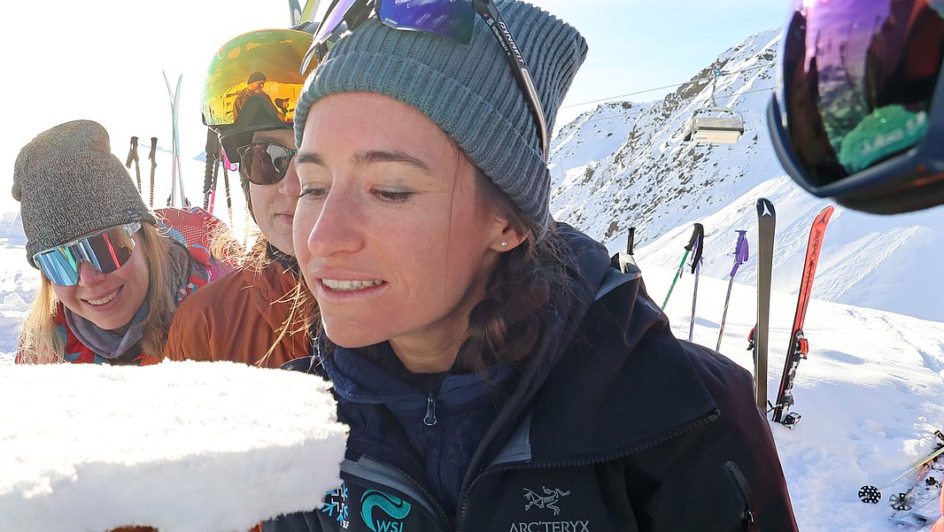 A group of four people in winter clothing is gathered in a snowy landscape. One person is holding a chunk of snow, while another leans in to examine it closely. Ski equipment is visible in the background, indicating a mountain or ski area setting.
