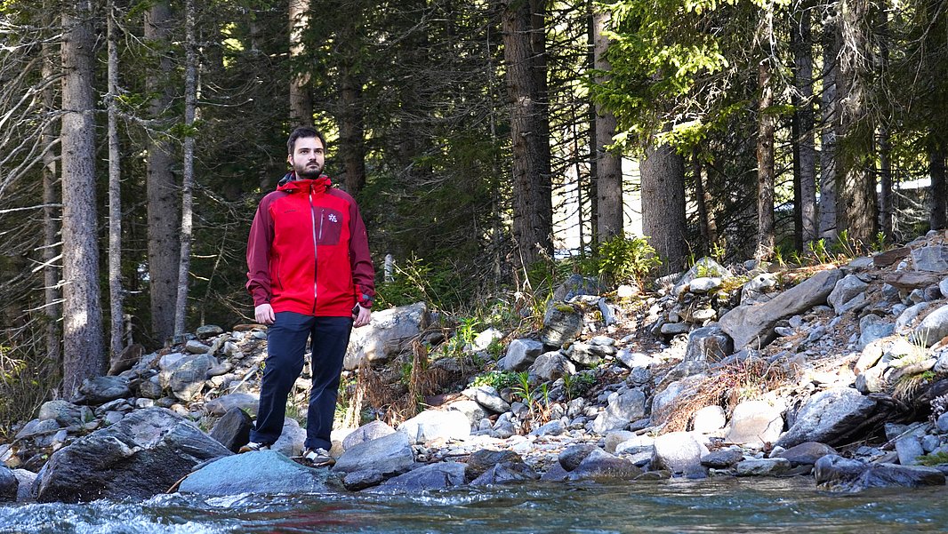Ein Mann steht in einem roten Jacket am Ufer eines Flusses, umgeben von Bäumen und Felsen. Das Wasser fließt ruhig im Vordergrund, während die Landschaft im Hintergrund grün und natürlich wirkt.