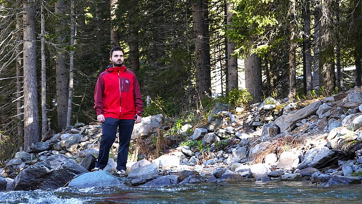 A person stands on a rocky riverbank, wearing a red jacket and navy pants. The background features tall trees and some greenery, with clear water flowing in front. Sunlight creates a bright atmosphere, highlighting the natural setting.