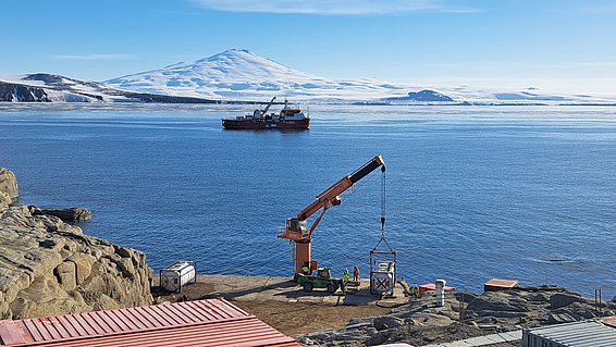 Im Vordergrund sieht man einen kleinen Hafenkran. In der Mitte des Bildes befindet sich ein ruhiges Meer mit einem mittelgrossen Schiff darauf, während im Hintergrund schneebedeckte Berge zu sehen sind.