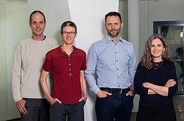 Vier Personen stehen nebeneinander in einem modernen Raum. Sie tragen legere Kleidung und lächeln in die Kamera. Der Hintergrund zeigt helle Wände und Elemente moderner Architektur.