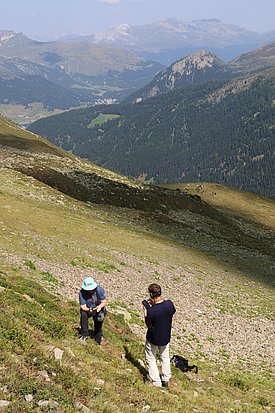 Frau mit helblauem Sonnenhut, blauem T-Shirt und schwarzer Hose und Mann mit dunklem T-Shirt und heller Hose an einem Berghang, im Hinergrund eine Bergkette, dazwischen ein Tal