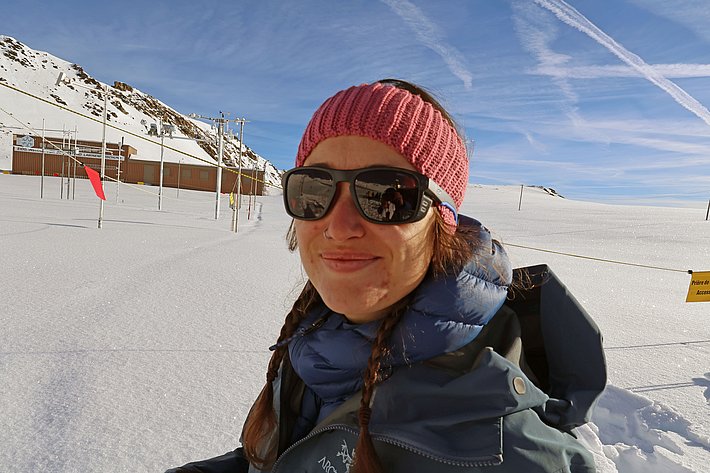 A person wearing sunglasses and a pink headband smiles in a snowy landscape. The background features snow-covered hills, and there are poles with flags in the distance, indicating a snowy environment suitable for winter activities.