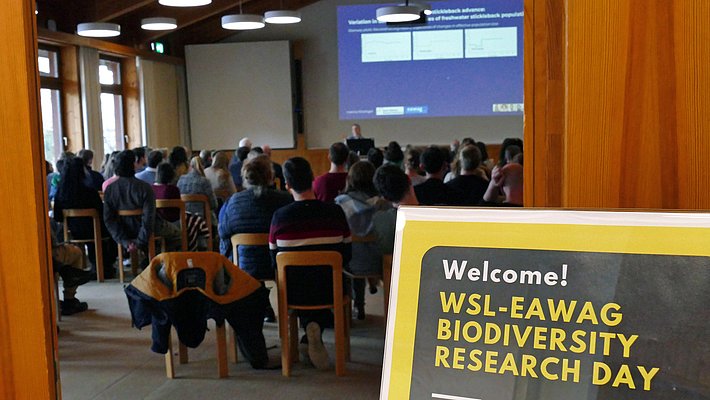 An audience is gathered in a lecture hall for the WSL-EAWAG Biodiversity Research Day. A sign in the foreground welcomes attendees, while a presentation is displayed on a screen in the background, indicating the event's focus on biodiversity research.