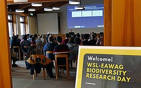 Ein Raum voller Teilnehmer bei einer Veranstaltung zum Thema Biodiversität. Im Vordergrund steht ein Willkommensschild, das auf den WSL-EAWAG Biodiversity Research Day hinweist. Die Präsentation im Hintergrund zeigt Informationen zu Forschungsprojekten.