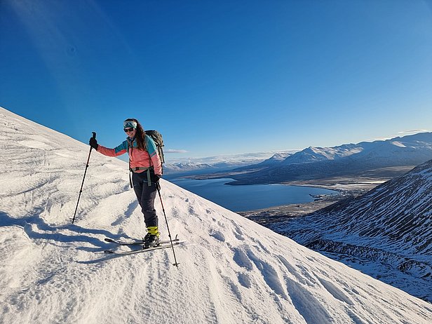 A skier stands on a snow-covered slope, using ski poles for support. The background features a vast landscape with mountains and a serene lake under a clear blue sky. The scene conveys an adventurous winter atmosphere.
