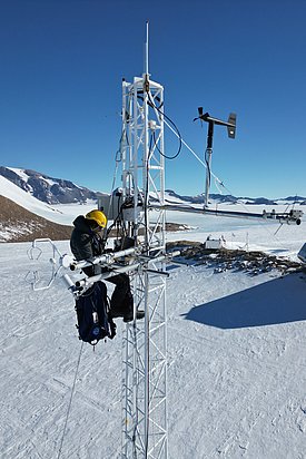 Person mit gelbem Helm arbeitet an einer Wetterstation auf einem schneebedeckten Berg mit Forschungsstation im Hintergrund
