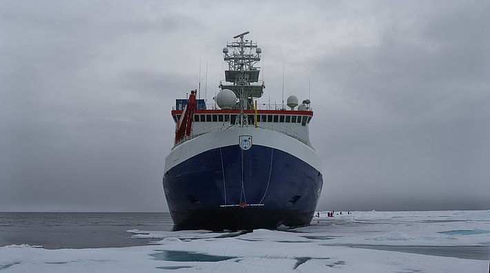 Ein großes Forschungsschiff mit blau-weißem Rumpf ist von vorne zu sehen, wie es in eisigen Gewässern mit verstreutem Meereis unter einem bewölkten Himmel vor Anker liegt.