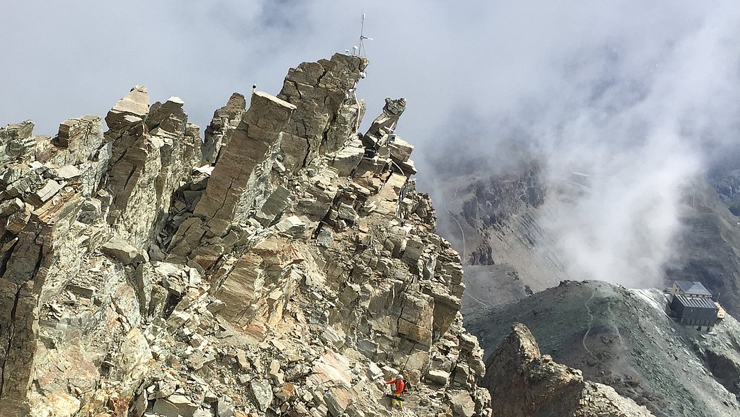 Cresta rocciosa con formazioni rocciose scoscese, circondata da nuvole e da un paesaggio montano sullo sfondo.