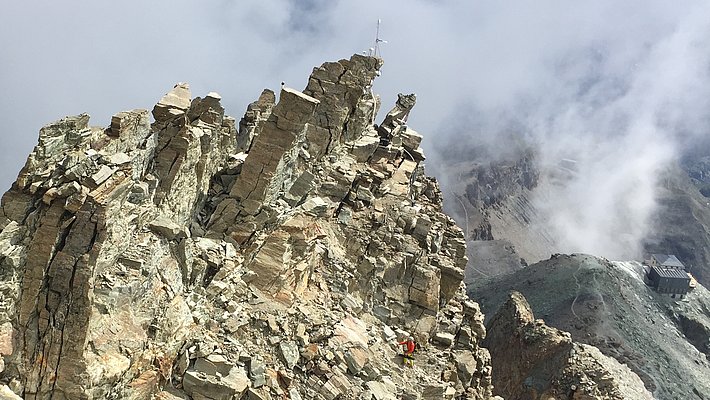 Crête avec des formations rocheuses escarpées, entourée de nuages et d'un paysage de montagne en arrière-plan.