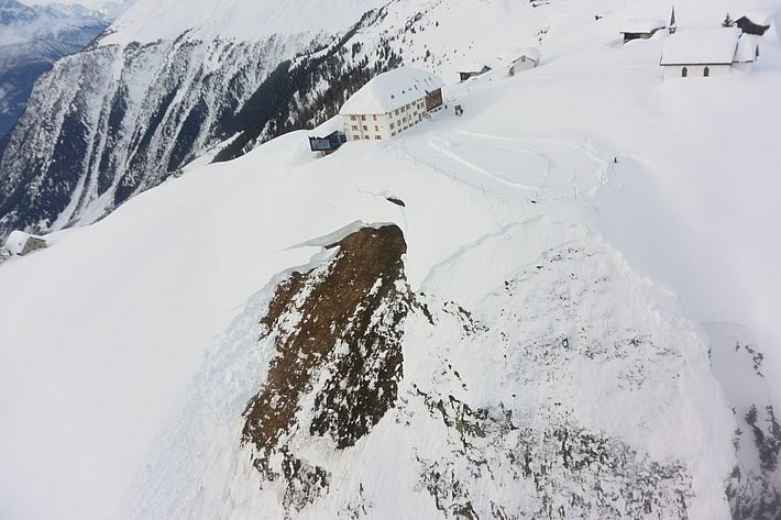 Das Bild zeigt das, auf einem Bergrücken gelegene Hotel Belalp. Circa fünfzig  Meter unterhalb des Hotels erstreckt sich der Anriss einer mächtigen Gleitschneelawine, die den darunterliegenden Boden freigelegt hat.
