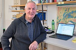 Ein Mann sitztan einem Tisch mit einem Laptop. Im Hintergrund sind Regale mit Ordnern und Büchern zu sehen. Der Raum wirkt akademisch, und auf dem Tisch liegt eine Karte. Der Mann trägt einen grauen Pullover und lächelt.