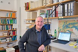 Un homme souriant est assis à un bureau, avec un ordinateur portable ouvert devant lui. En arrière-plan, des étagères remplies de livres et de dossiers sont visibles. La pièce semble être un bureau de travail.