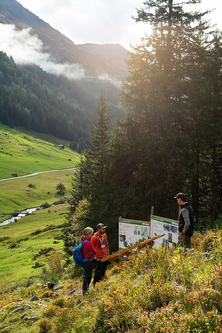 Drei Personen stehen vor zwei Informationsschildern in einer bergigen Landschaft. Im Hintergrund sind grüne Wiesen und Bäume sichtbar, mit einem Fluss, der durch das Tal fliesst.
