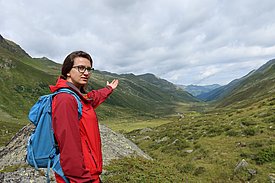 A woman with a red jacket and blue rucksack points to a green valley surrounded by mountains under a cloudy sky.