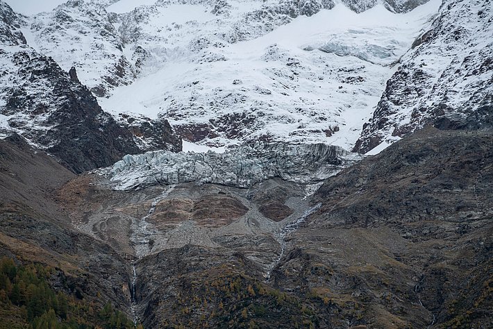 Glacier on a snow-covered mountain with rocky terrain and scattered trees in the foreground.