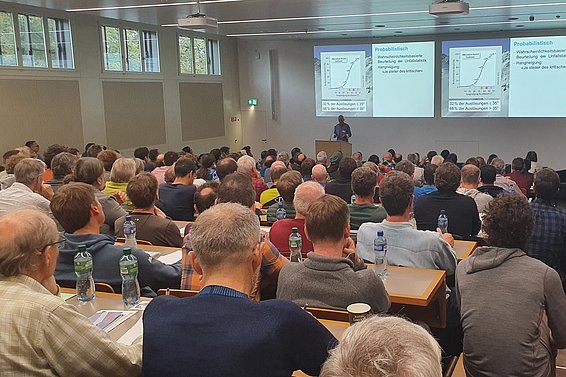 Ein Saal mit vielen Menschen drin; von hinten sieht man nach vorne, wo jemand einen Vortrag hält.