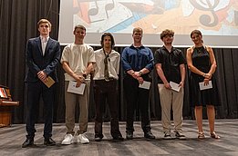 Un groupe de six jeunes gens se tient en ligne sur scène lors d'un événement. Ils portent des tenues variées, allant de formelles à décontractées. Chaque personne tient un document dans les mains, et un écran en arrière-plan présente des éléments graphiques liés à la musique.