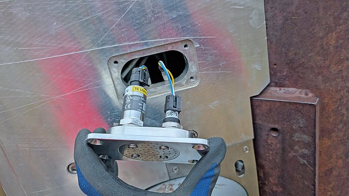 A close-up view of a person's hand in a glove holding a metal component featuring two sensors with wires, positioned near a large, gray metallic surface with a rectangular opening.