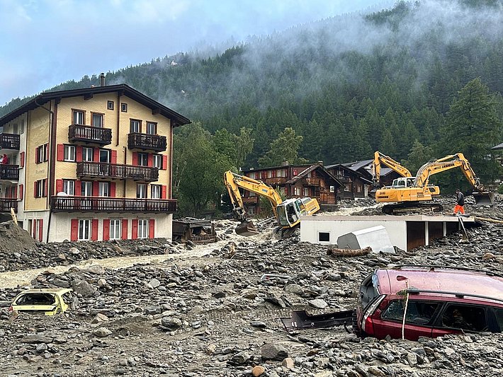 Zwei gelbe Bagger arbeiten auf einem felsigen Gelände vor einem mehrstöckigen Haus mit roten Fensterläden, daneben liegt ein beschädigtes rotes Auto, im Hintergrund bewaldete Berge mit Nebel.