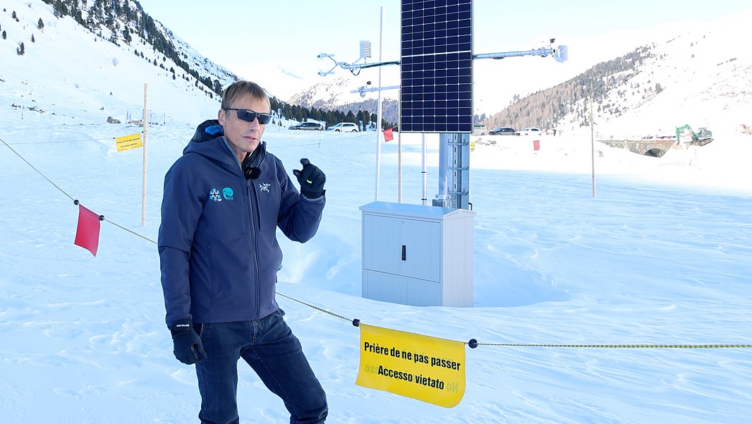 Person mit Sonnenbrille und Winterjacke steht neben einer Messstation mit Solarpanel in verschneiter Berglandschaft
