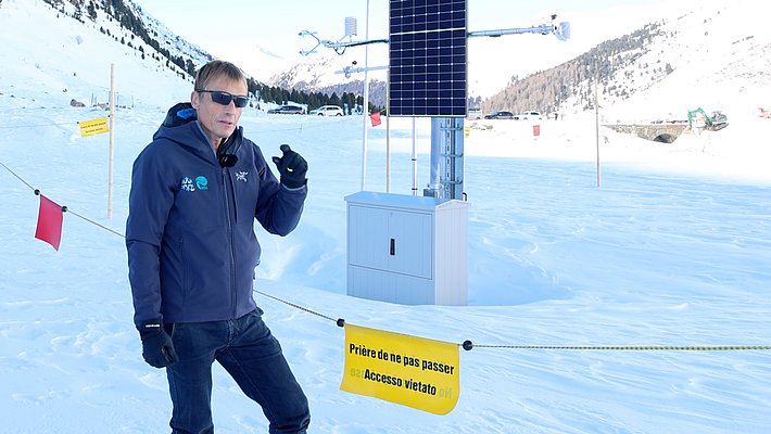 Person wearing sunglasses and winter jacket standing next to a measurement station with solar panel in snowy mountain landscape