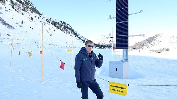 Person mit Sonnenbrille und Winterjacke steht neben einer Messstation mit Solarpanel in verschneiter Berglandschaft