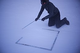 Person in dunkler Winterkleidung kniet im Schnee und misst mit Metalllinealen ein Quadrat auf dem Boden.