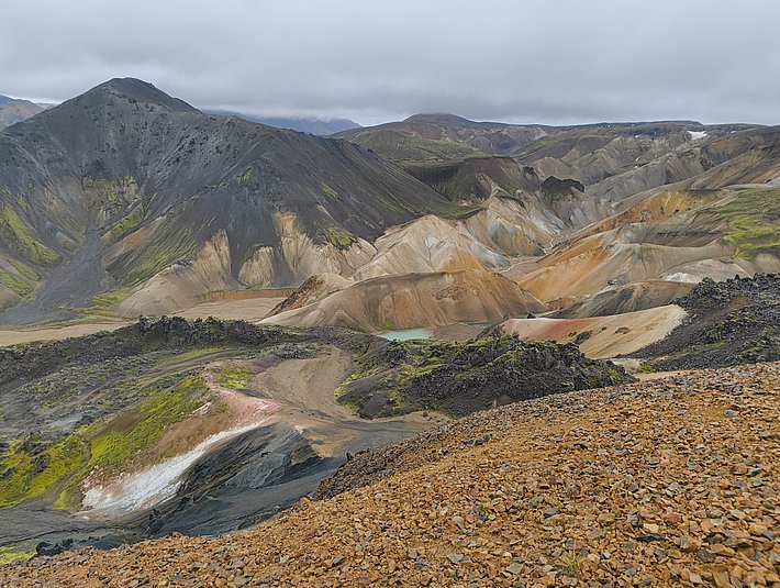 A panoramic view of a rugged volcanic landscape featuring multicolored mountains and textured soil. Shades of brown, black, and hints of green contrast with patches of reddish hues, creating a striking natural scene under a cloudy sky.