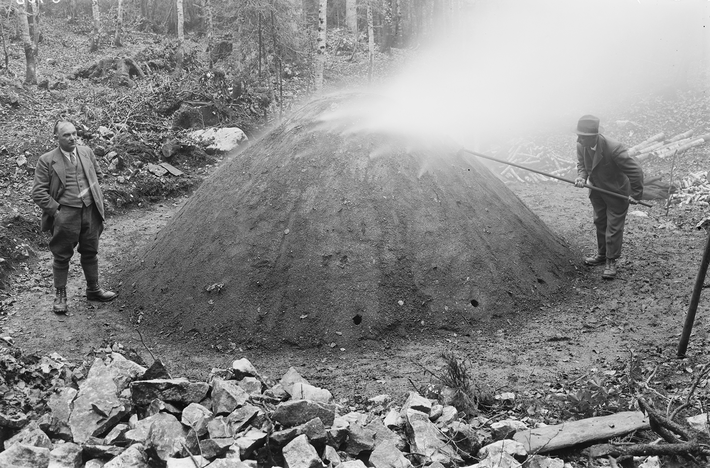 Zwei Männer stehen in einem Waldgebiet neben einem großen, Hügel. Einer der Männer stockert mit einem langen Stock, im Hügel, aus dem Rauch aufsteigt.