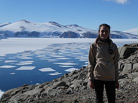 Anja lächelt in die Kamera. Hinter ihr sind ein mit Eis bedecktes Meer und Schneeberge sichtbar, der Himmel ist strahlend blau.