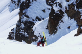 Person mit roter Hose und blauem Rucksack trägt gelbe Skier an einem steilen, schneebedeckten Berghang mit Felsen.