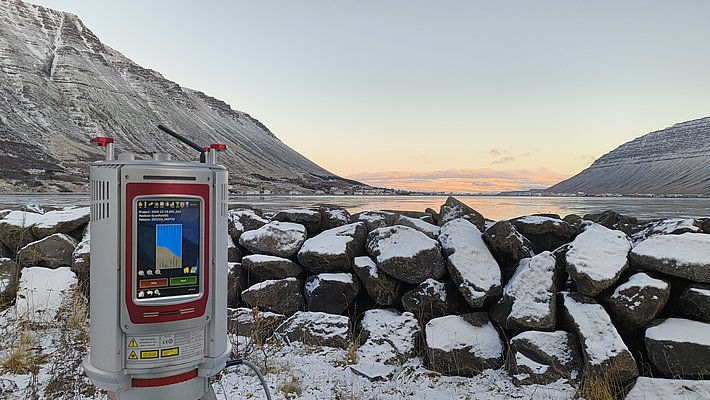 L'image montre un appareil de mesure sur un trépied, situé devant un paysage de montagnes enneigées et d'un lac tranquille. La scène est baignée dans une lumière douce du matin, avec des rochers en arrière-plan et un ciel dégagé.