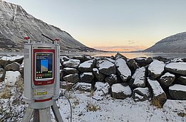A surveying instrument is positioned on a tripod in a snowy landscape, with a river and rocky terrain in the background. The scene features mountains rising on either side and a clear sky, indicating early morning or late afternoon light.