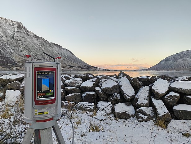 A surveying instrument is positioned on a tripod in a snowy landscape, with a river and rocky terrain in the background. The scene features mountains rising on either side and a clear sky, indicating early morning or late afternoon light.
