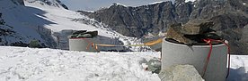 Auf einem schneebedeckten Hang am Matterhorn sind zwei Betonrohre senkrecht in den Schnee und den darunterliegenden Permafrost gegraben. Ihre Metalldeckel sind mit grossen Steinen beschwert, die beiden Rohre sind mit einer losen Leine verbunden. Im Hintergrund der Bohrlöcher erstreckt sich ein Gletscherabbruch und dahinter eine schroffe Felswand.